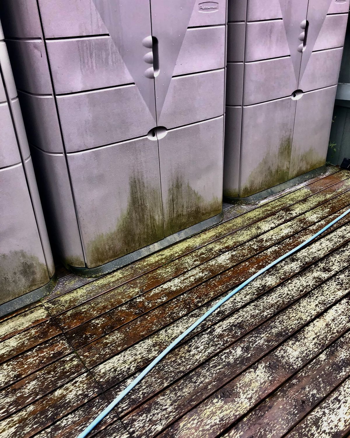 A weathered wooden deck streaked with mildew, green staining up the base of two plastic storage cabinets