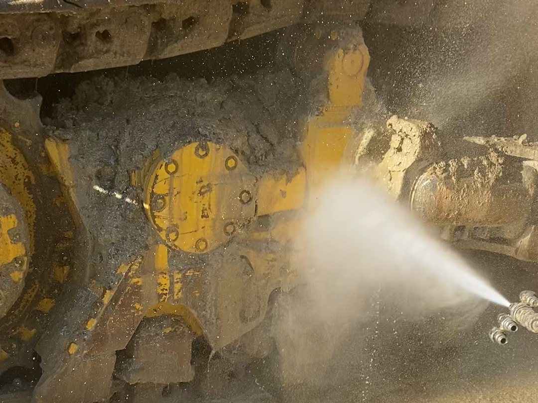 Pressure washing packed mud out of a bulldozer's track frame, the spray cutting back to yellow paint
