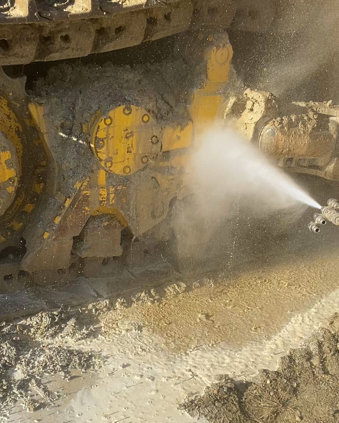 Pressure washing packed mud out of a bulldozer's track frame, grey slurry pooling on the ground below