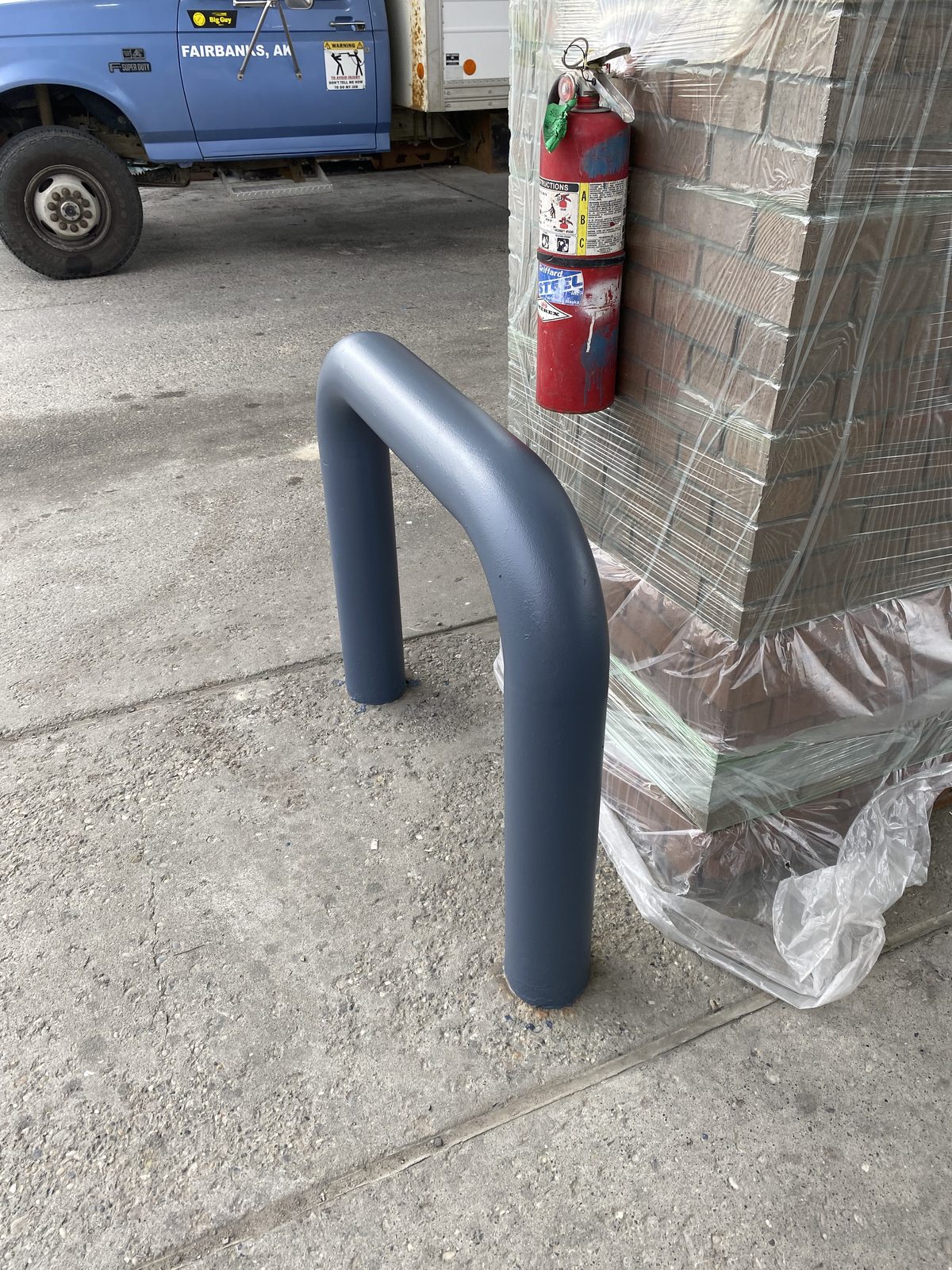 A steel bollard freshly painted blue-grey beside a brick column still wrapped in plastic