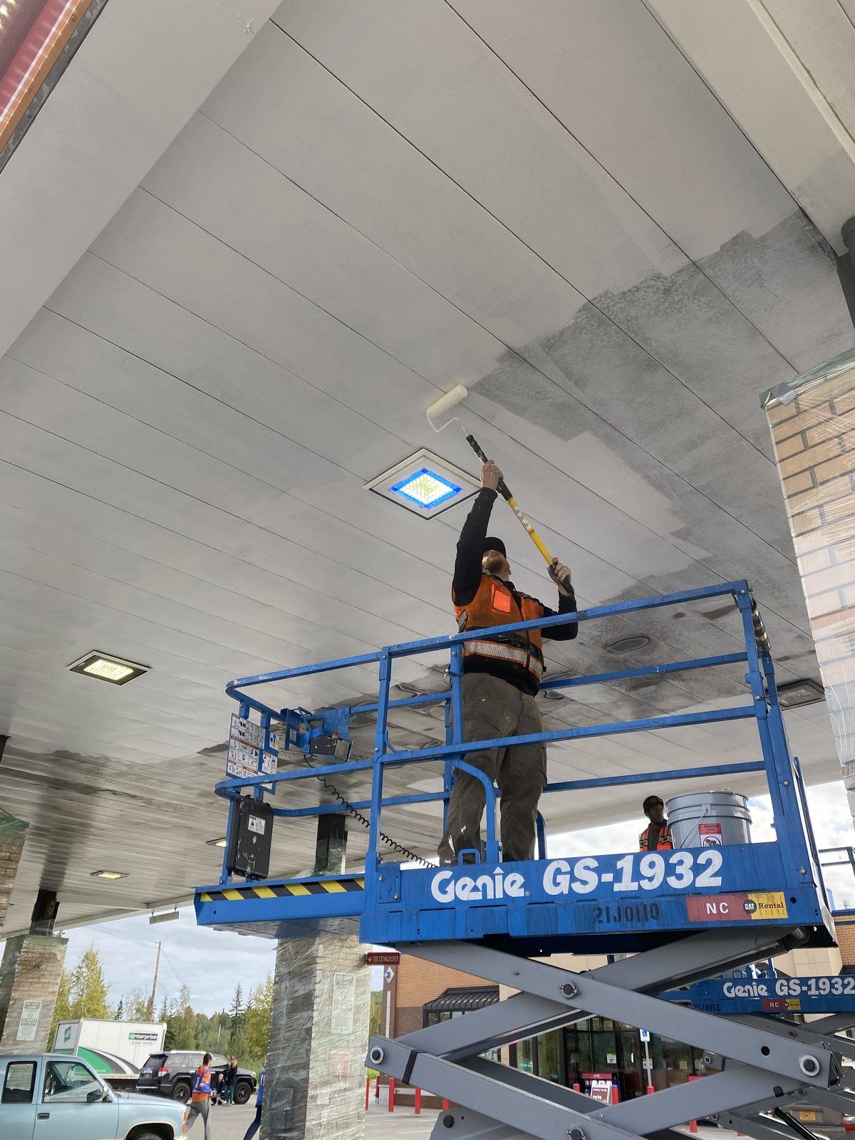 Rolling fresh paint onto the canopy ceiling from a scissor lift, the painted half bright against the grey