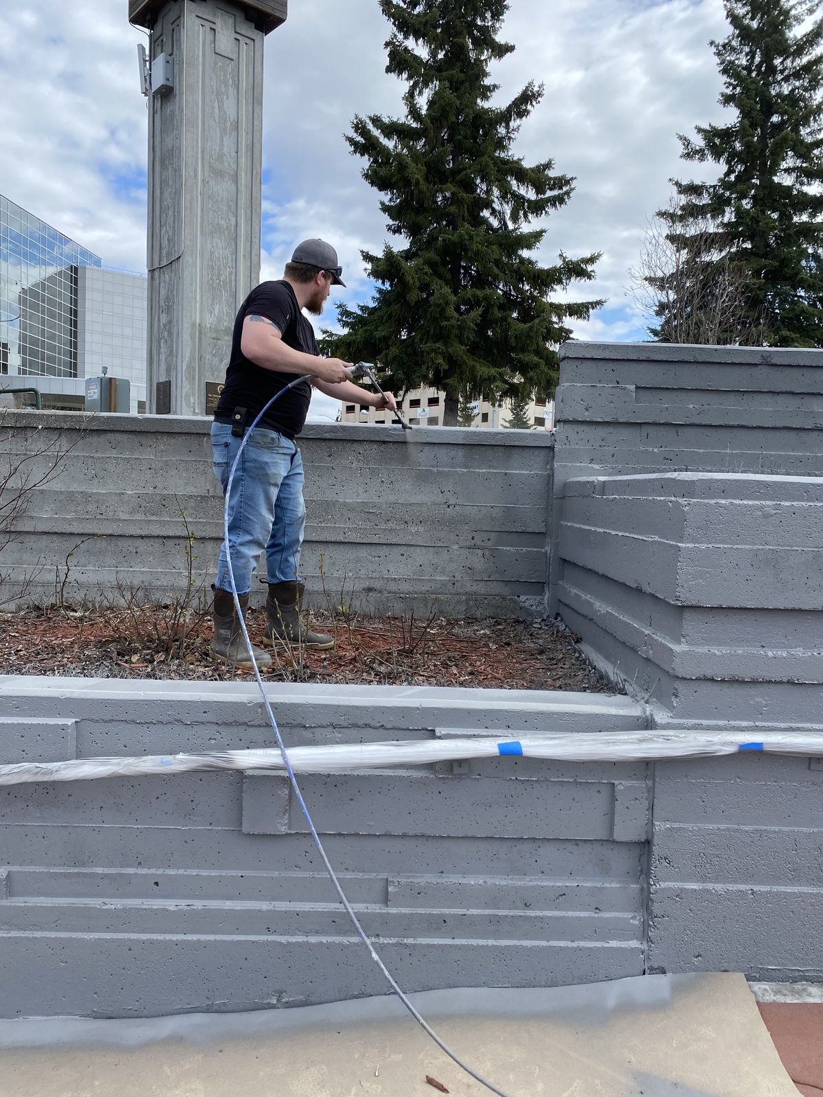Spraying grey paint onto the plaza walls, plastic sheeting laid along the base to protect the pavers