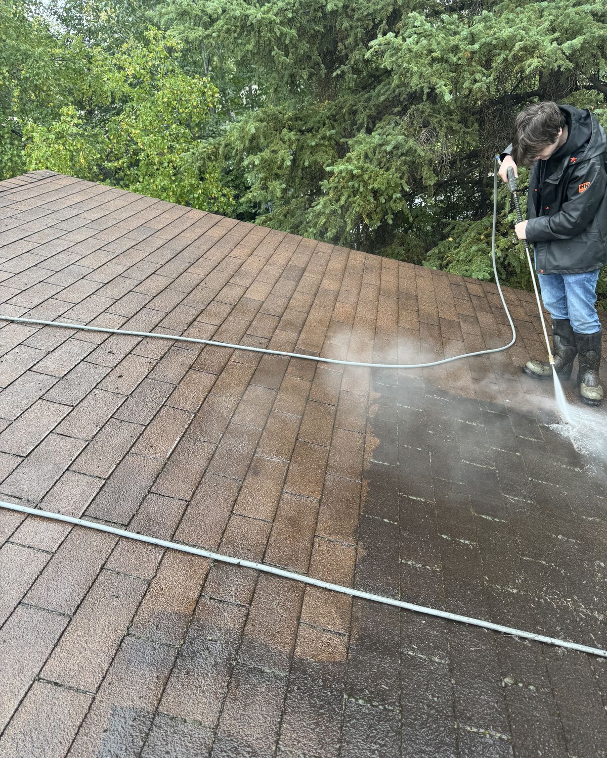 Joel's son Timothy in rain gear pressure washing the shingle roof, a clear line between the washed shingles and the dirty ones