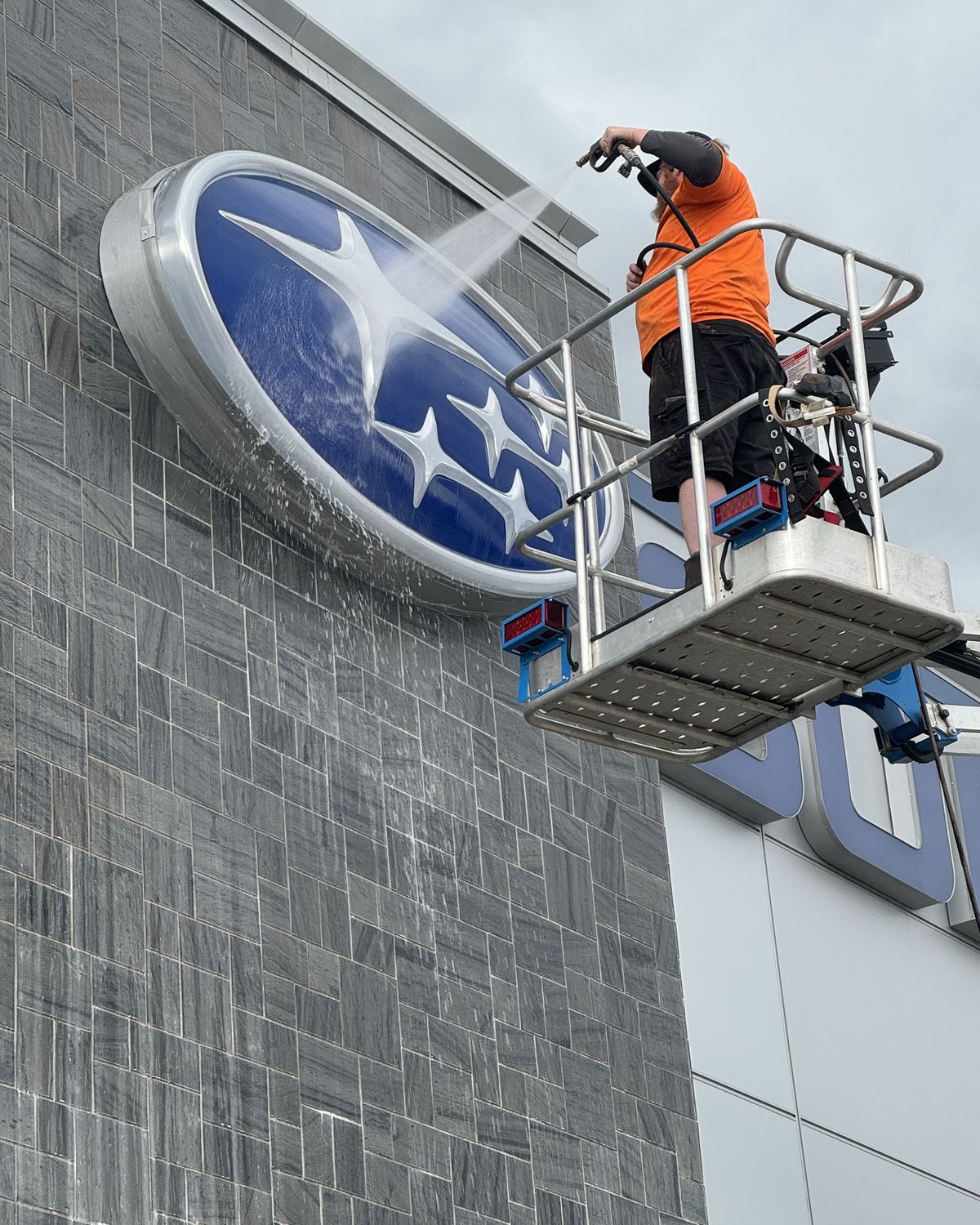 Joel's nephew Adam washing a dealership's wall-mounted sign from a lift basket, spray hitting the sign face